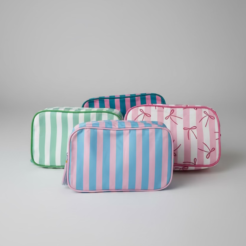 Three rectangular zippered pouches with striped patterns are arranged on a white quilted blanket. One pouch has pink and blue vertical stripes, one has green and white stripes, and one has pink and white vertical stripes with a bow print.