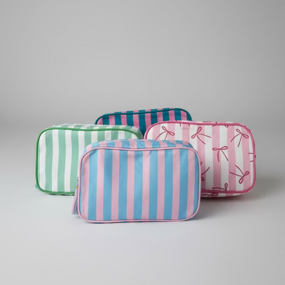 Three rectangular zippered pouches with striped patterns are arranged on a white quilted blanket. One pouch has pink and blue vertical stripes, one has green and white stripes, and one has pink and white vertical stripes with a bow print.